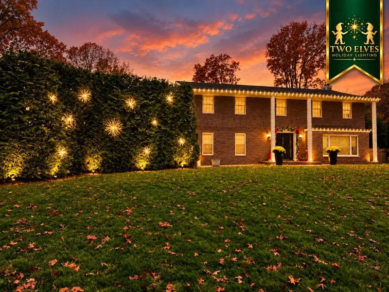 Brick colonial at sunset with roofline lights and starburst-lit hedge